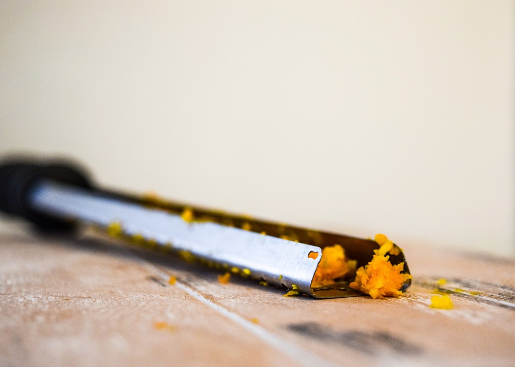 a zester laying upside down on a pale white washed wooden plank with orange zest inside the zester