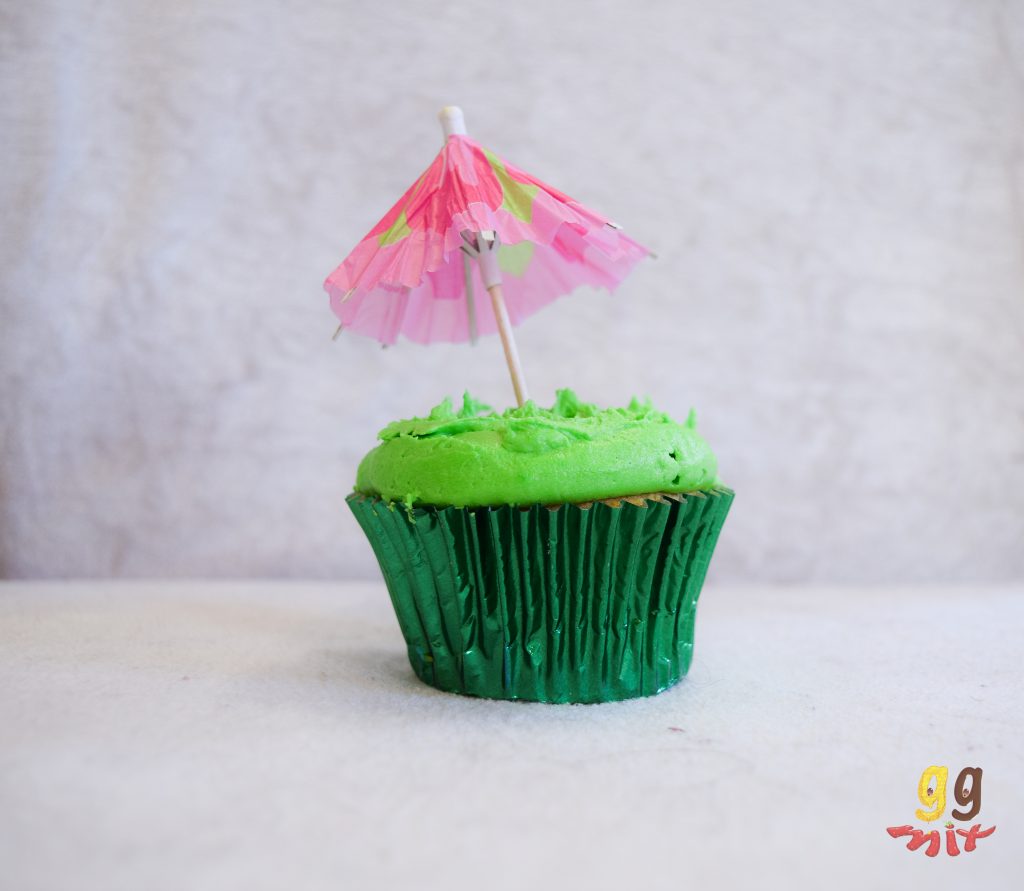 a cupecake in a metalic green cupcake case with lime green buttercream on the top and a pink cocktail umbrella in the middle of the cupcake