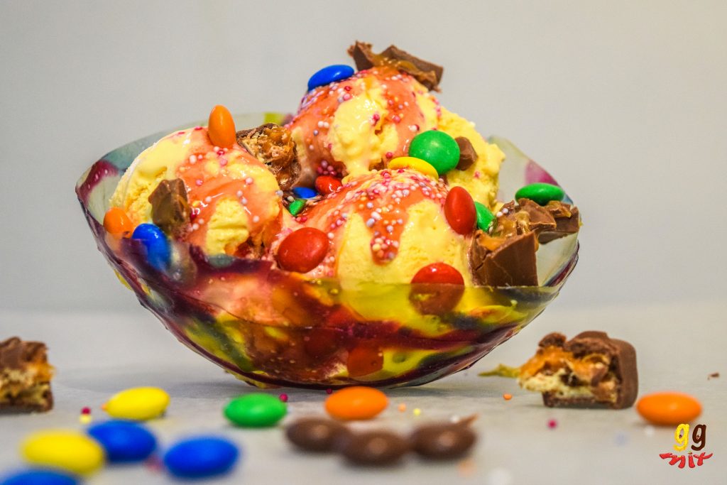 a multi coloured gelatin bowl filled with ice cream, smarties, chocolate pieces, strawberry sauce and sprinkles