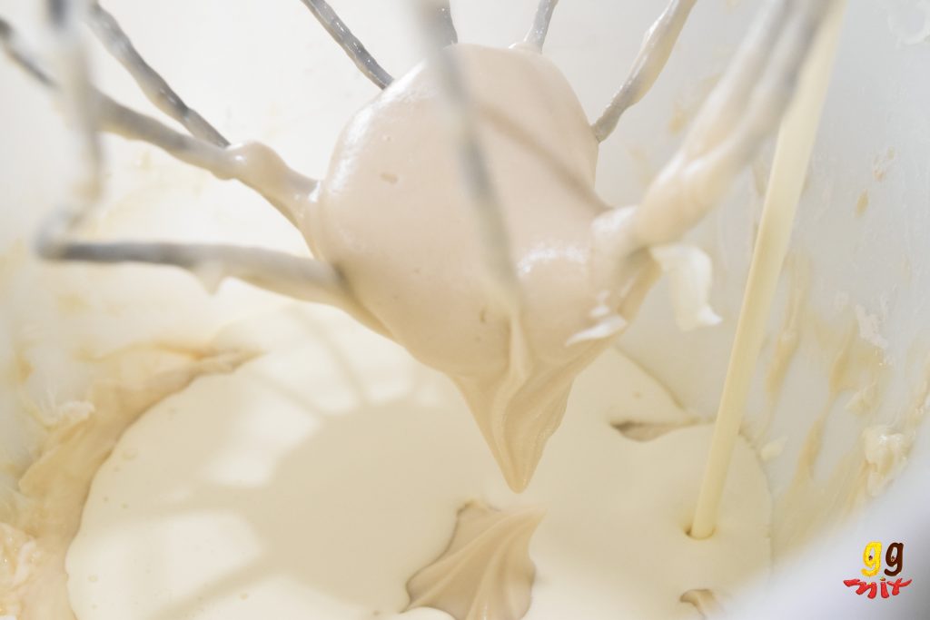 a bowl of cream cheese frosting, a standmixer whisk and cream being poured in