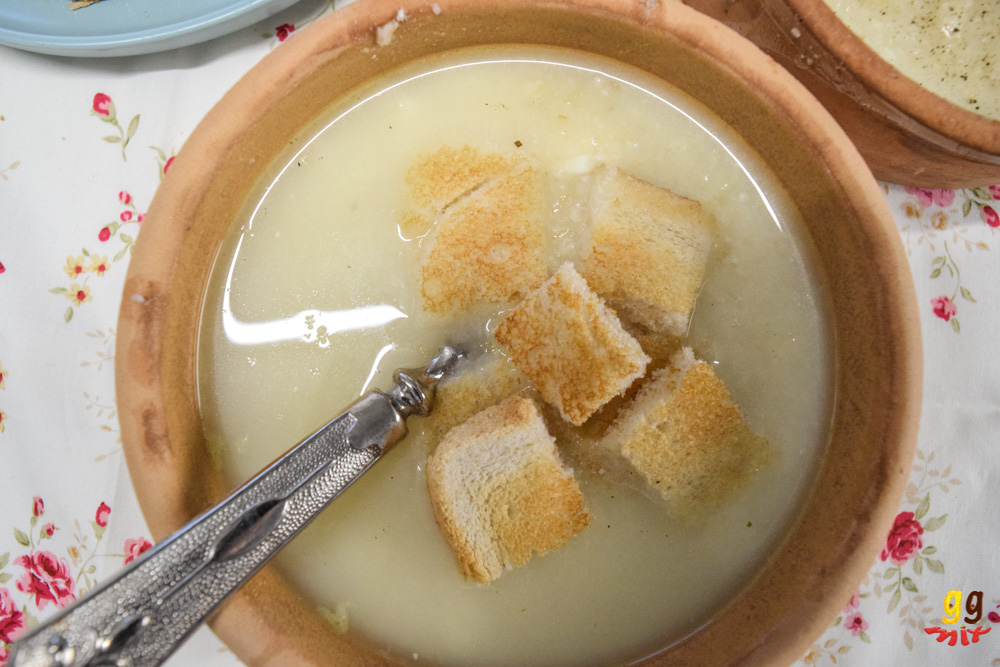 a brown clay bowl filled with trahanas me halloumi soup topped with cubes of toast and a traditional spoon Cypriot spoon, sitting on a white pink rose tablecloth