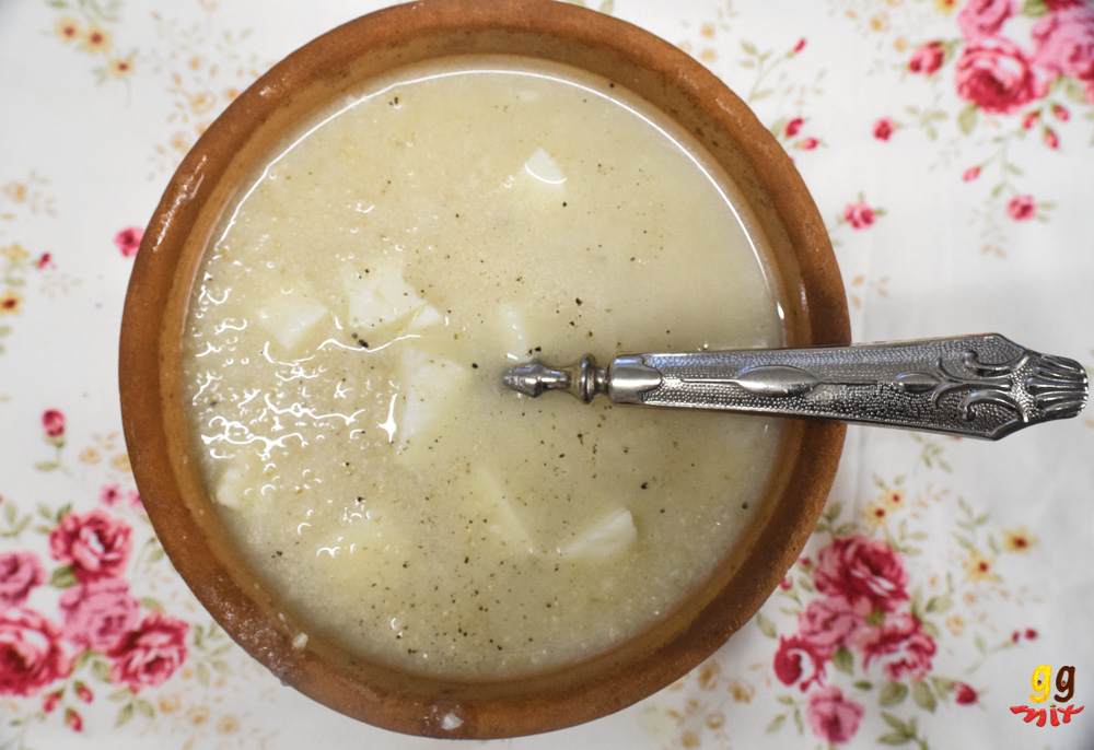 a brown clay bowl filled with trahanas me halloumi soup with a sprinkling of black pepper and a traditional spoon Cypriot spoon, sitting on a white pink rose tablecloth
